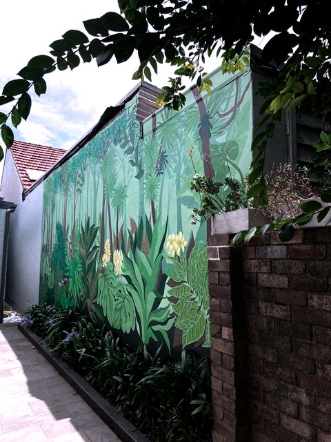 Mural de selva tropical en tonos verdes cubriendo una pared exterior junto a un jardín