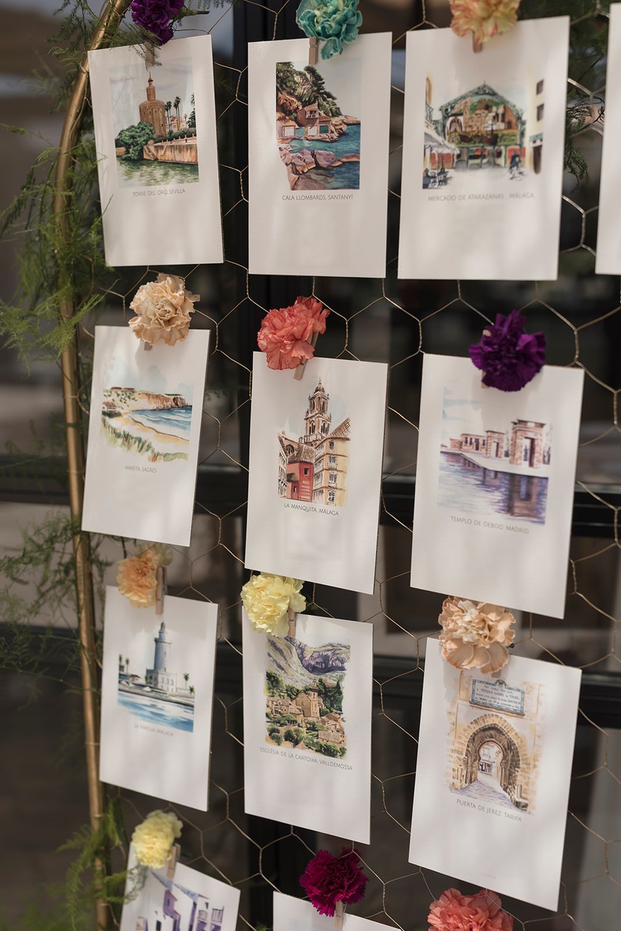 Seating plan de boda con una acuarela de un lugar distinto en cada mesa, colgado sobre malla con claveles