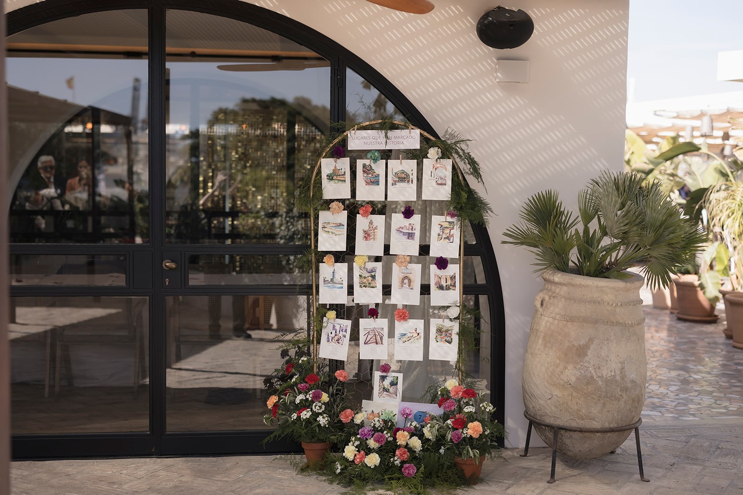 Seating plan de boda pintado en acuarela, con una lámina por mesa colgada en un arco con flores a la entrada del banquete